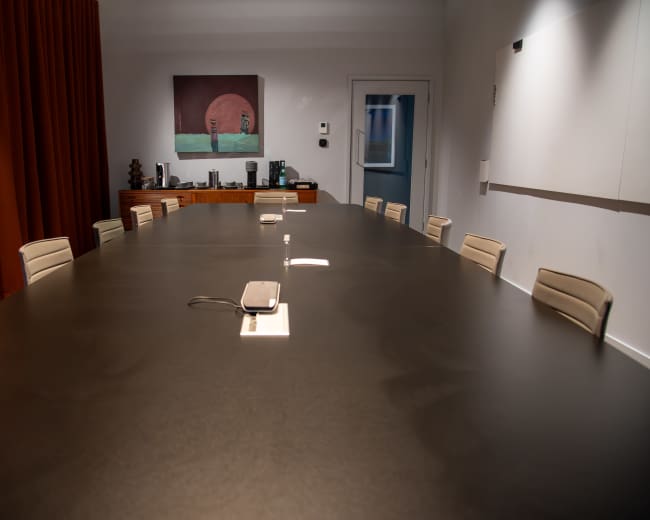 A large conference room features a long black table surrounded by beige chairs, with a painting on the wall and equipment visibly arranged in the background.