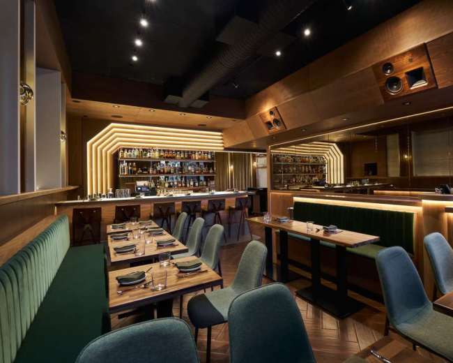 The image shows a modern restaurant interior featuring wooden furnishings, green upholstered seating, and ambient lighting highlighting a bar area.