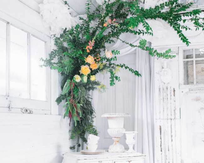 A white wooden cabinet stands in a room adorned with greenery and flowers, complemented by light-colored curtains and rustic decor.