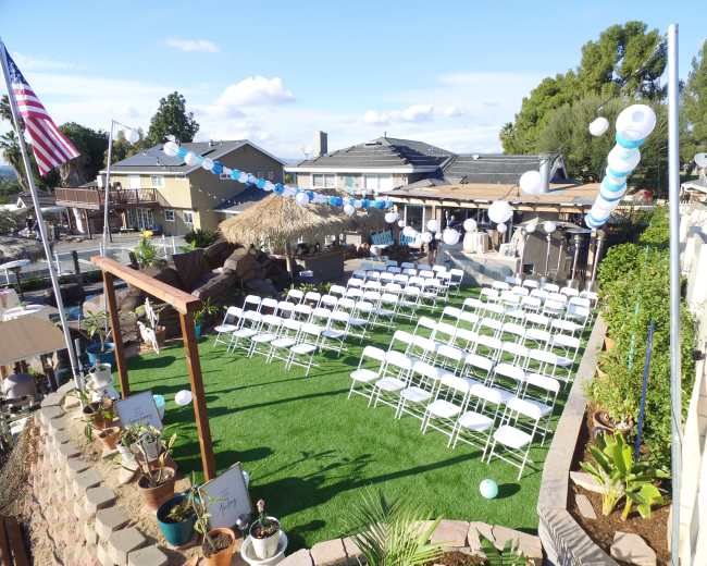 A neatly arranged outdoor seating area with white chairs set up on green grass, surrounded by decorative balloons and a thatched-roof structure.