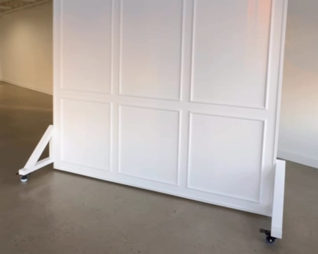 The image shows a large, white, freestanding panel with a simple, segmented design, placed on a smooth floor in an empty room.