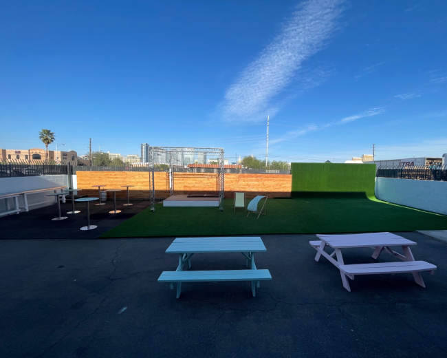 The image shows an outdoor venue with a green turf area, picnic tables, a wooden stage, and a backdrop of a blue sky.