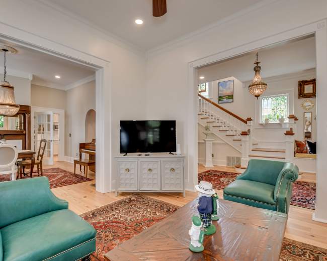 A bright, open living space features two teal armchairs, a wooden coffee table, and a television, with a staircase and another room visible in the background.