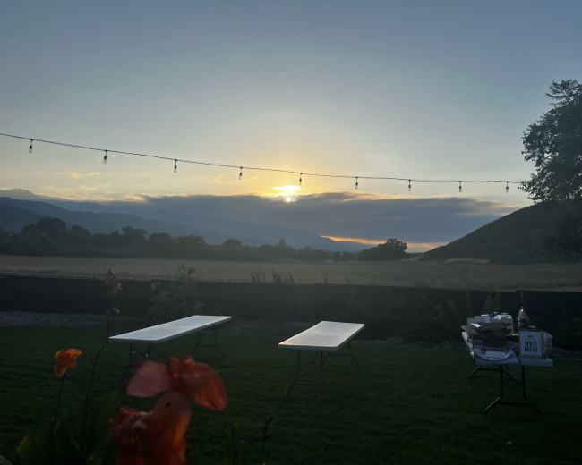 The scene features a sunset over distant mountains, with tables set up on a grassy area and string lights overhead.