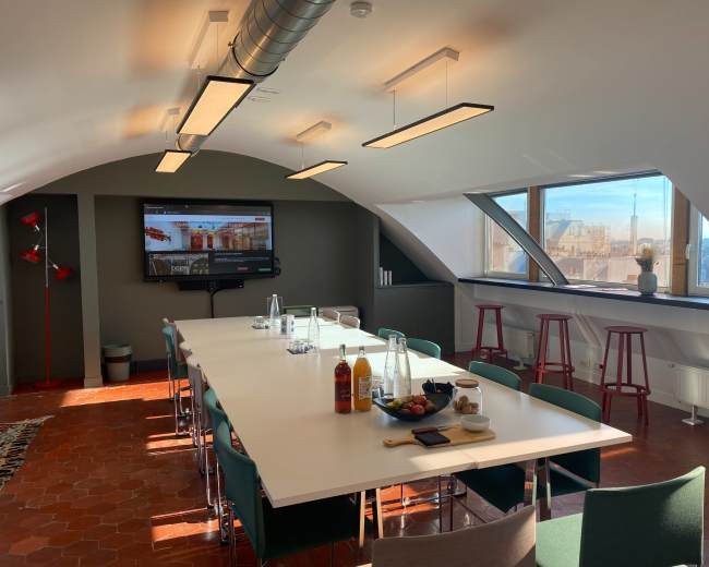 A modern conference room features a long white table, colorful chairs, and large windows with a city view, displaying a wall-mounted screen.