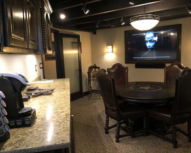 The image shows a kitchen area with a granite countertop, a coffee maker, and a wooden dining table surrounded by chairs, with a television mounted on the wall.
