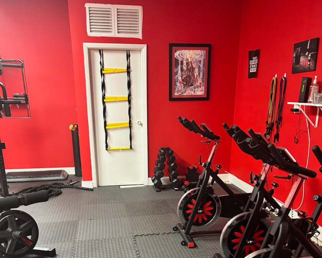 The image shows a gym area with black exercise equipment, including stationary bikes and weights, set against red walls and a patterned rubber floor.