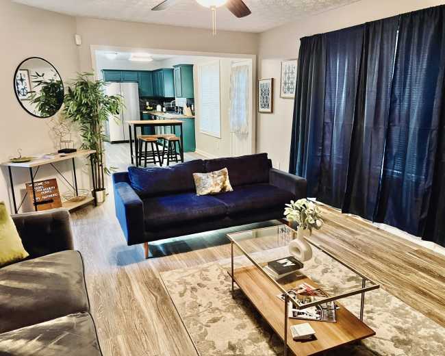 The image shows a cozy living room with a dark blue sofa, a glass coffee table on a patterned rug, and a doorway leading to a kitchen with teal cabinets.
