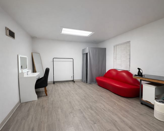 The image shows a minimalist room featuring a red seating area, a dressing mirror, a clothing rack, and a gray privacy curtain.