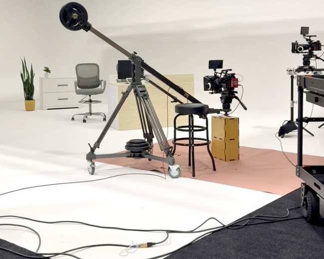 A film studio setup features a camera on a dolly track, a chair, a desk, and various filming equipment in a minimalistic white space.