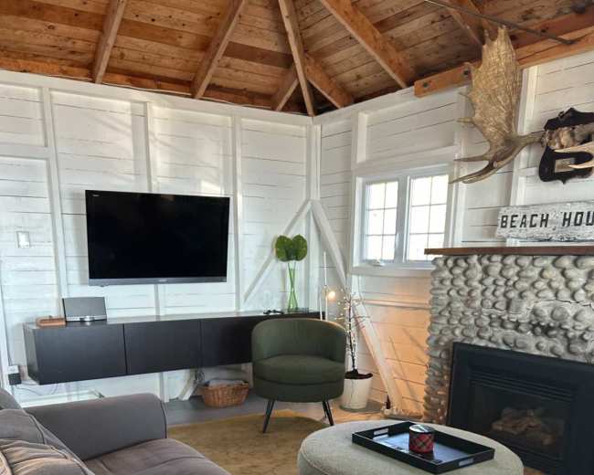The image shows a cozy living room with a gray sectional sofa, a circular coffee table, and a mounted flat-screen TV against a white wooden wall with a stone fireplace.