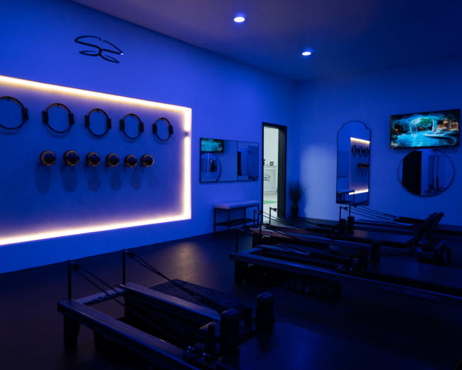 A fitness studio features reformer machines arranged neatly under a blue-lit wall with circular wall decorations and a television displaying a pool scene.
