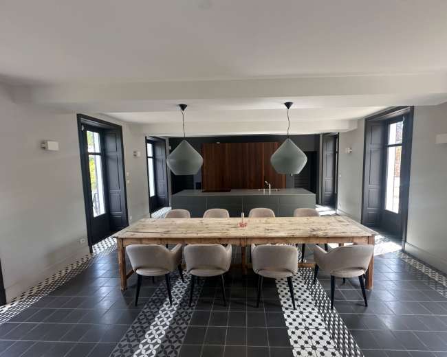 A large wooden dining table with eight chairs is set in a modern room featuring gray walls, pendant lighting, and tiled flooring with a geometric pattern.