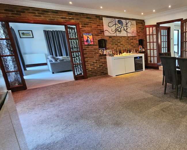 The image shows a spacious interior area with a dining table and chairs, adjacent to a bar setup, featuring brick walls and a carpeted floor.