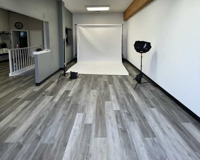 A spacious photography studio with a plain white backdrop and studio lighting set up against a light-colored wooden ceiling and gray flooring.