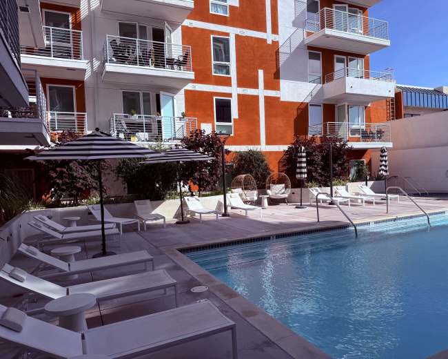 The image shows a modern outdoor pool area surrounded by lounge chairs, a striped umbrella, and colorful apartment buildings in the background.