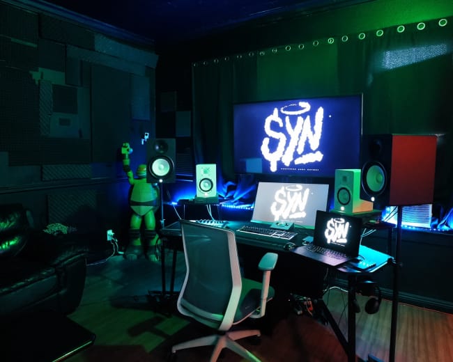The image shows a modern music studio with a desk, computer, speakers, and colorful lighting.