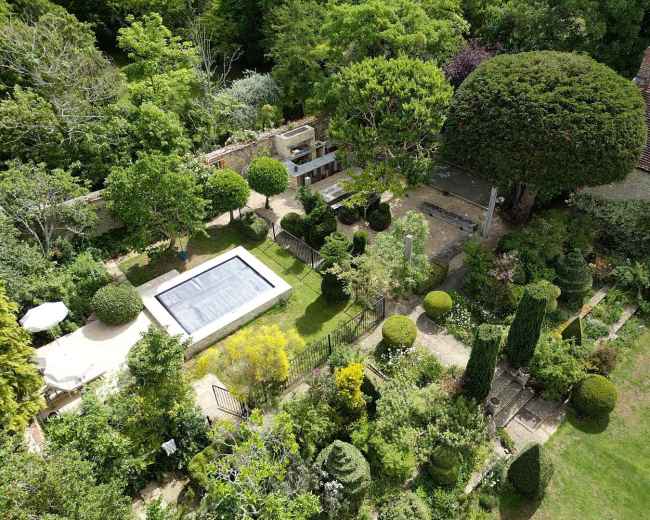 The image shows a landscaped garden with a swimming pool, various plants, and neatly trimmed hedges surrounded by trees.