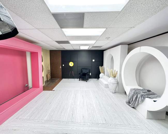The image shows a modern studio space with a black accent wall, a large pink frame, a white circular structure, and a chair with decorative elements arranged neatly.
