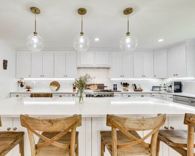 A modern kitchen features white cabinets, a large island with wooden stools, and pendant lighting above.