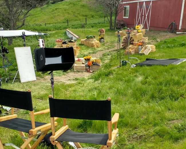 A film set in a grassy area with director's chairs facing a scene decorated with hay bales and flowers near a red barn.