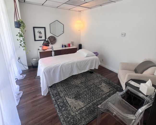 The image depicts a treatment room with a massage table covered in white linens, a cozy chair, and decorative elements on the walls.