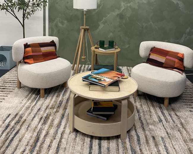 The image shows a cozy seating area with two rounded chairs, a wooden coffee table stacked with books, a floor lamp, and a green patterned wall in the background.
