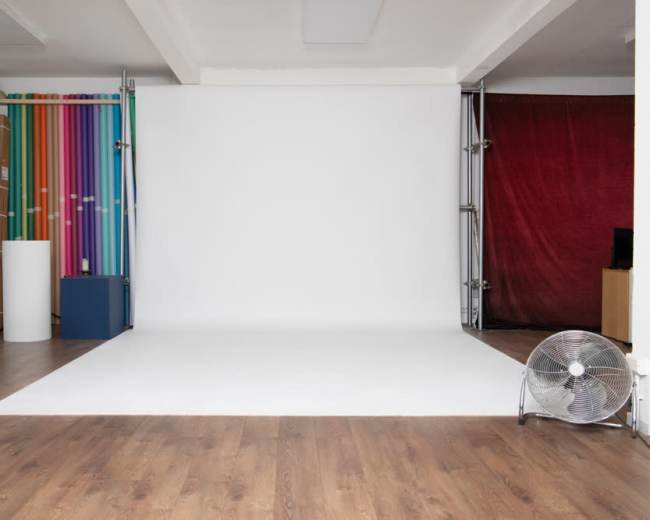 The image shows a photography studio with a blank white backdrop and colorful striped rolls on one side, along with a fan and wooden flooring.