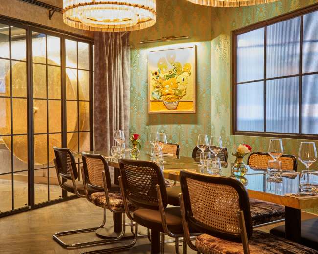 The image shows a modern dining room featuring a large table surrounded by chairs, with decorative wallpaper and pendant lighting, and a glass partition revealing a large circular feature.