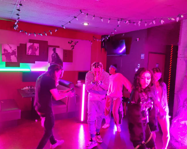 A group of people dances and interacts inside a brightly lit room with neon lights and a vibrant atmosphere.