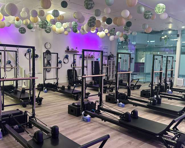 The image shows a brightly lit fitness studio with several reformer Pilates machines set up in rows beneath decorative paper lanterns hanging from the ceiling.