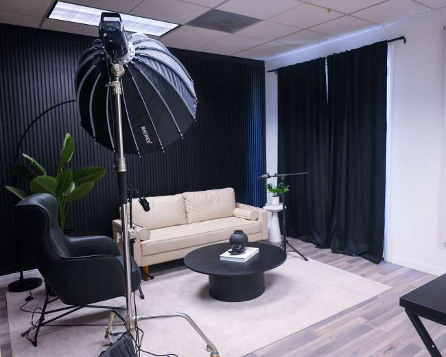 The image features a modern studio setup with a cream-colored sofa, a round black coffee table, and various plants, all arranged in a well-lit room with dark curtains and a backdrop.