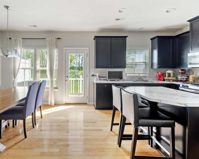 A modern kitchen and dining area features a wooden table with chairs, a kitchen island with bar stools, and large windows overlooking a view.