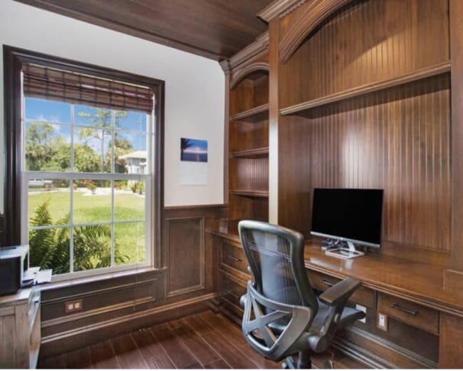 The image shows a wooden home office with a large desk, an ergonomic chair, and a computer monitor facing a window with natural light.