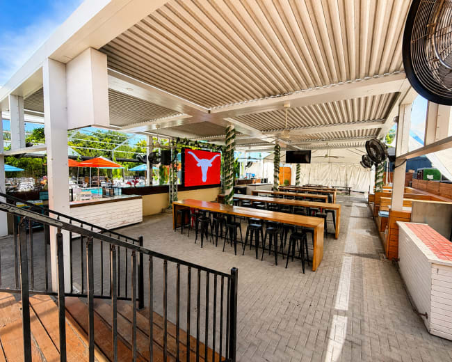The image shows a spacious outdoor dining area with long wooden tables, black chairs, and a large red flag featuring a white symbol hanging on the wall.