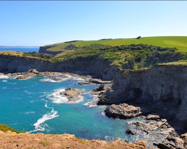 A coastline features rocky cliffs and clear blue waters, with green hills rising in the background.