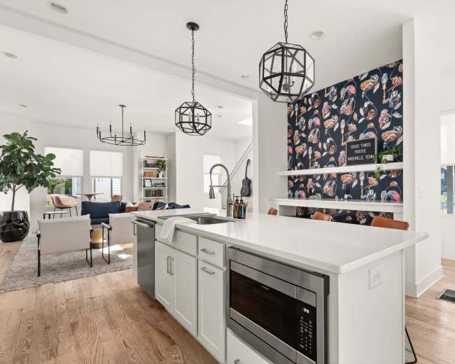 The image shows a modern kitchen with a white countertop, dark cabinetry, colorful wallpaper featuring food illustrations, and a view of an open living area.