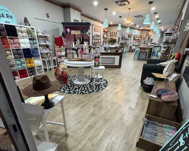 The image shows a cozy craft store interior with shelves stocked with yarn, a seating area, and various crafting supplies displayed throughout.
