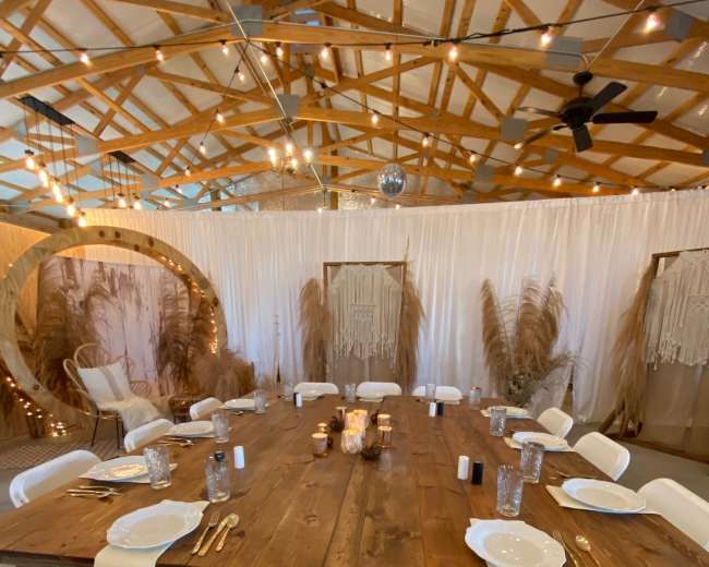 A long wooden table is set with white plates and glasses, surrounded by white folding chairs, in a decorated indoor space featuring string lights and decorative backdrops.