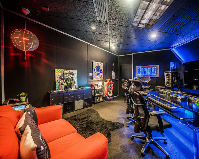 The image shows a modern music studio featuring a black wall, a computer desk with audio equipment, a red sofa, and vibrant decorative elements.