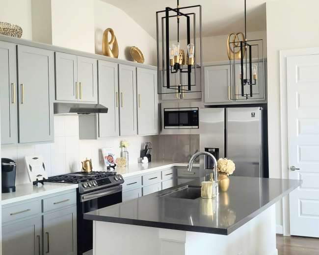 The image shows a modern kitchen featuring gray cabinets, stainless steel appliances, a center island with a dark countertop, and pendant lighting overhead.