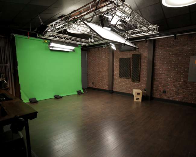 The image shows a video production studio featuring a green screen, dim lighting, and a wood-paneled floor.