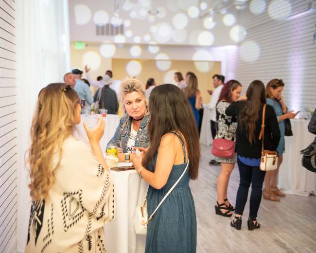 Two women converse at a gathering in a well-lit venue while others mingle in the background.
