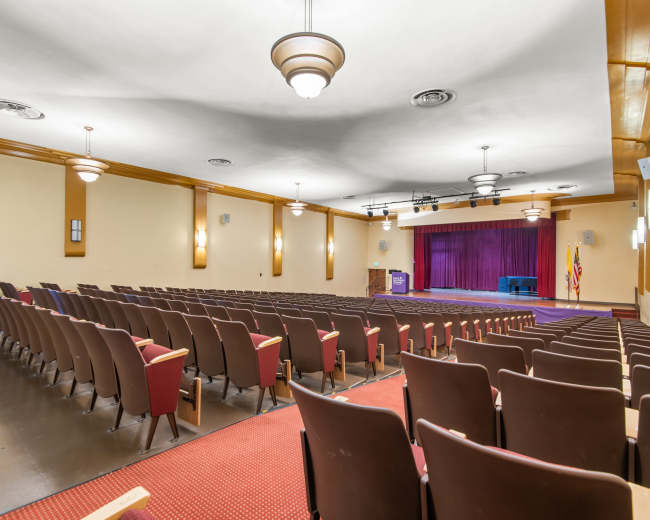 The image shows an empty auditorium with rows of brown chairs facing a purple stage, flanked by flags and equipped with stage lighting.