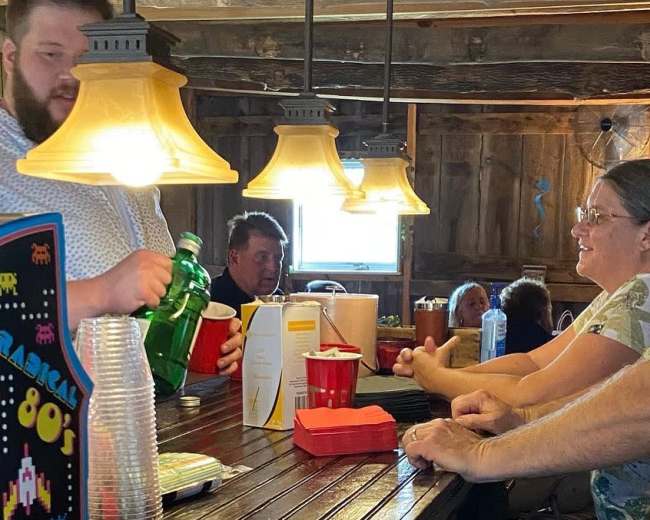 A bartender pours a drink for a patron at a rustic wooden bar, while several customers engage in conversation nearby.