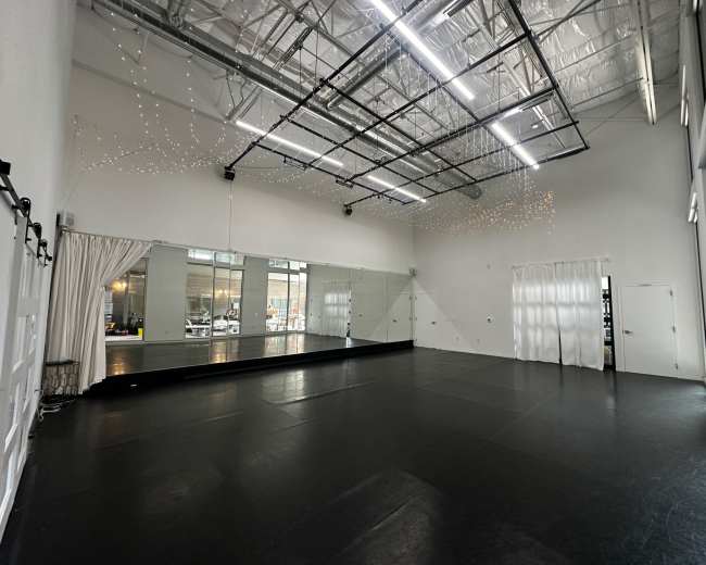 The image shows an empty dance studio with a black floor, large mirrors on one wall, and ceiling lights.