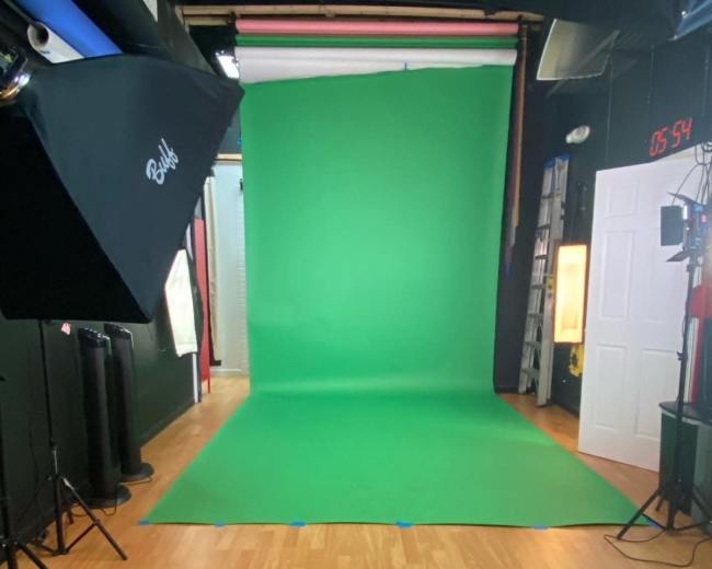 The image shows a photography studio featuring a green screen backdrop, professional lighting equipment, and a wooden floor.