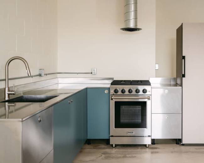 The image shows a modern kitchen with stainless steel countertops, an oven, a sink, and a refrigerator.
