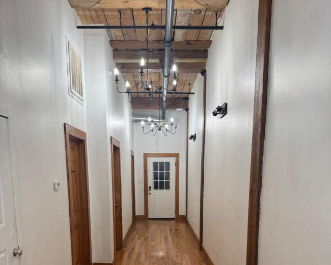 The image shows a long, narrow hallway with wooden flooring, white walls, and a door at the end, illuminated by overhead lights.