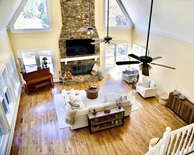 The image shows a spacious living room featuring large windows, a stone fireplace, a flat-screen TV, and a mix of furniture including a piano, sofas, and a coffee table.